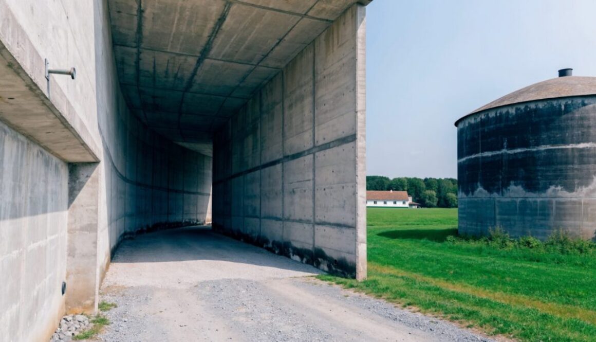 Fahrsilo mit hohen Sichtbeton-Wänden und runder Beton-Güllebehälter auf einem Bauernhof, grüne Wiese im Hintergrund