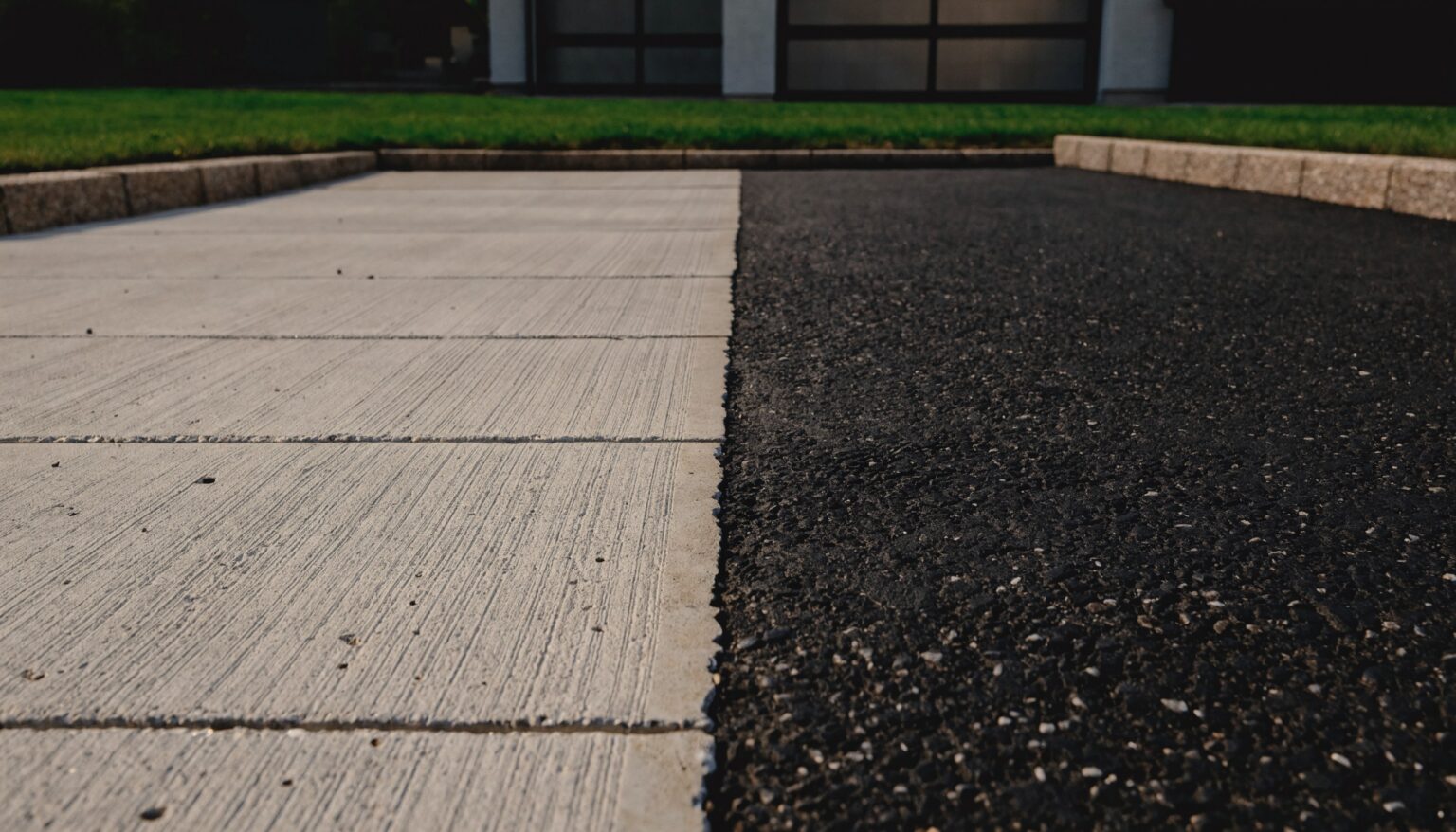 Asphaltieren oder betonieren: Einfahrt mit Betonfläche im Besenstrich neben einer Asphaltfläche, Randsteine und Garage im Hintergrund