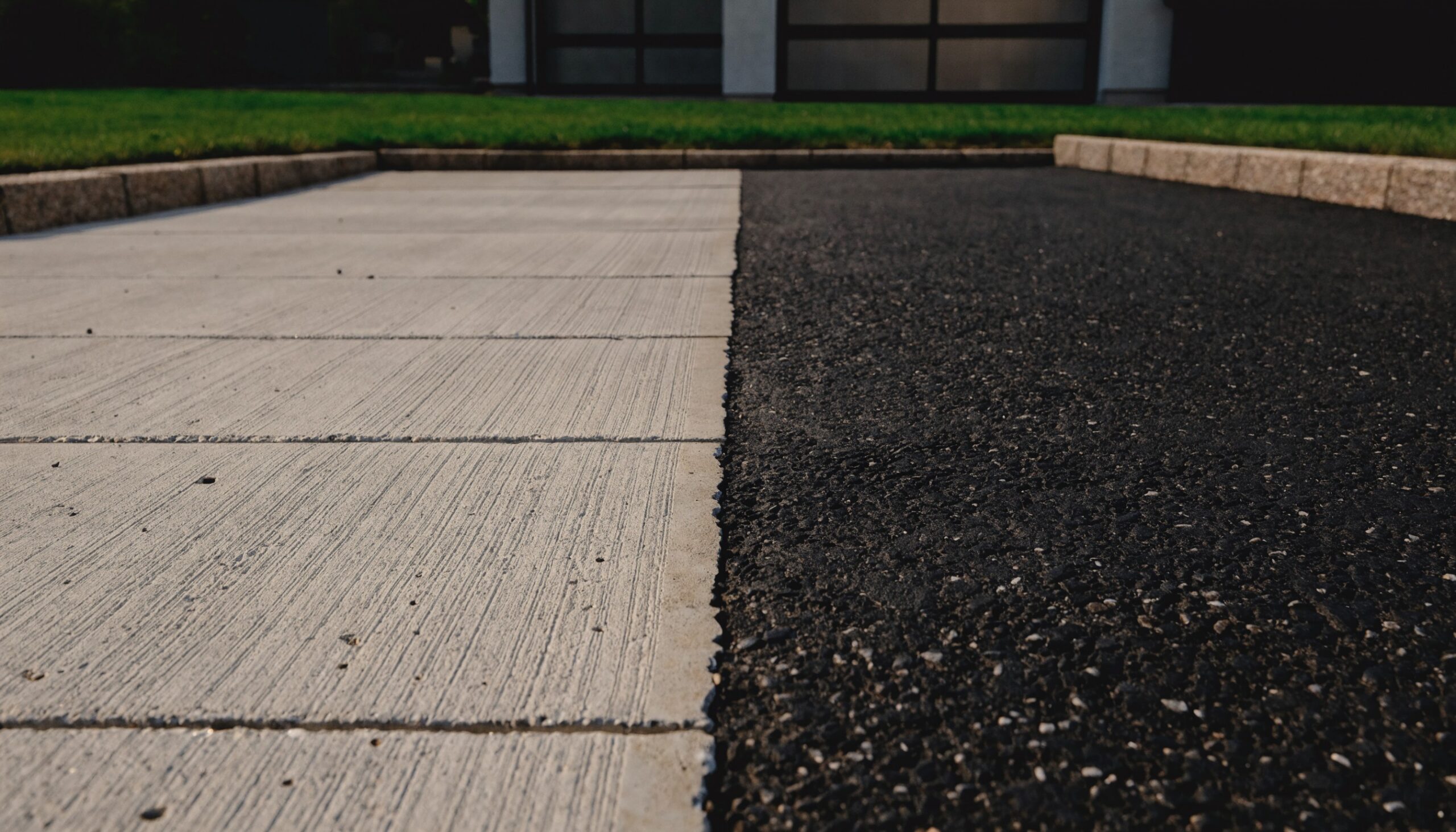 Asphaltieren oder betonieren: Einfahrt mit Betonfläche im Besenstrich neben einer Asphaltfläche, Randsteine und Garage im Hintergrund